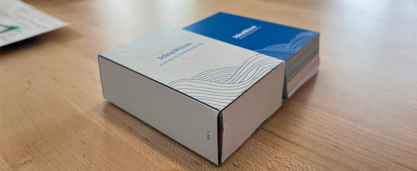 Two IdeaWave card boxes on a wooden table, one silver-white and one blue, printed with a wave pattern and the IdeaWave logo