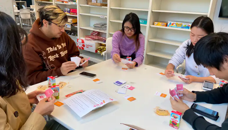 Graduate students at a white table holding cards and reading a printed instruction sheet, with snacks and phones on the table
