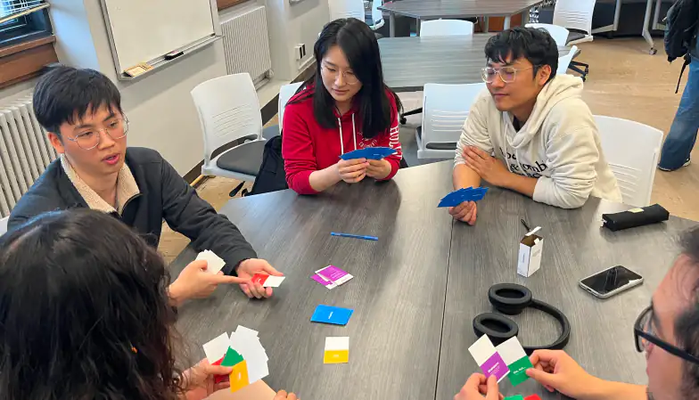 A graduate student gesturing as he explains an idea to peers who hold blue, pink and green cards around a gray table