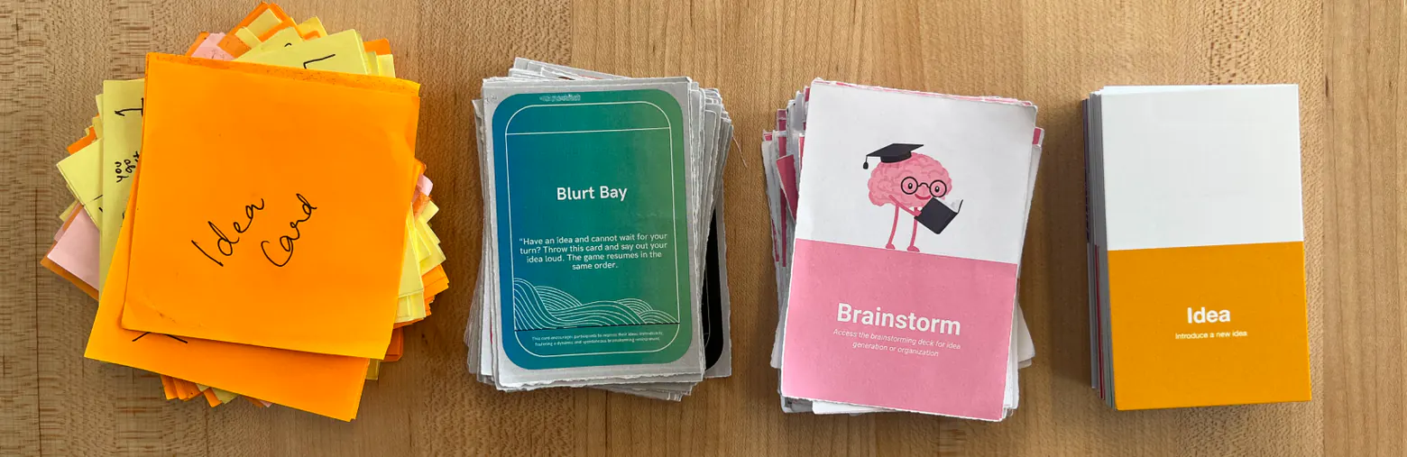 Four prototype generations on a wooden table: handwritten sticky-note cards, a teal “Blurt Bay” deck, a pink “Brainstorm” deck, and a polished white box with yellow cards