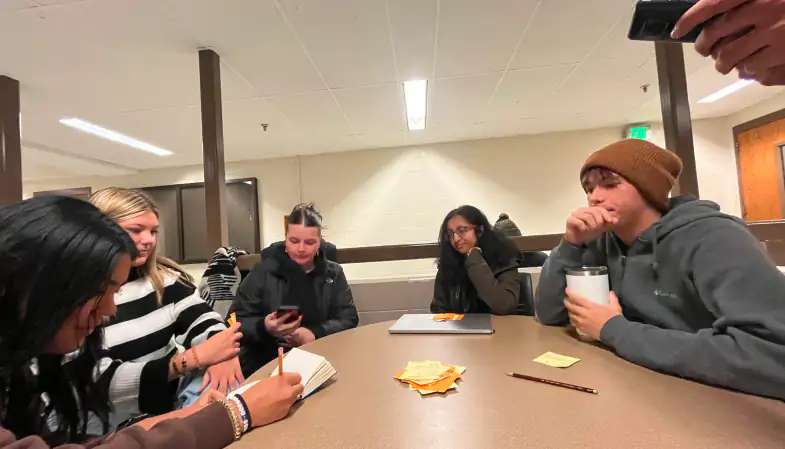 Five design students seated around a round table brainstorming with cards, sticky notes and notebooks during a testing session