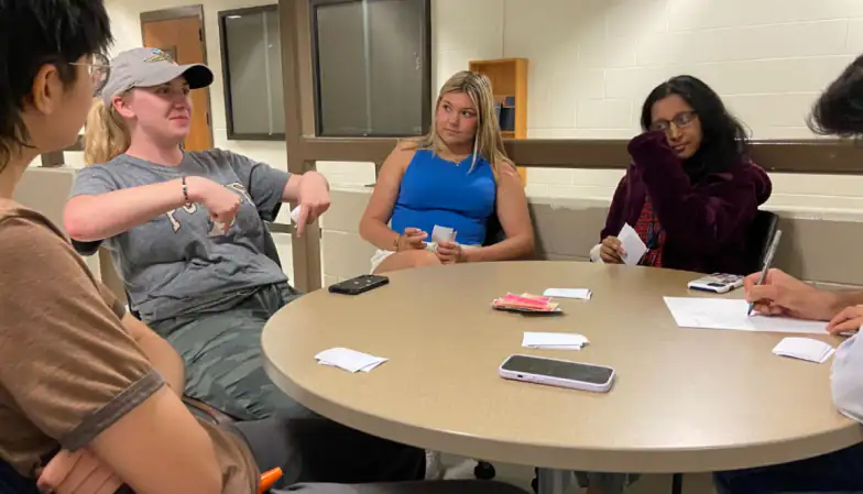 Students seated around a round table during a playtest, with cards and a phone on the table while the researcher writes notes on paper