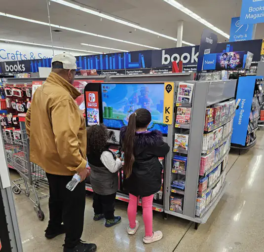 Two children and an adult watching a video-game display next to the book section of a Walmart