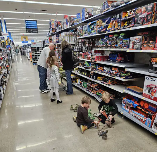 Two children sitting on the floor playing with toys in a Walmart toy aisle while two adults browse the shelves